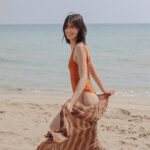 A model walking on the beach wearing an orange one piece swimsuit and a tie dye cotton brown stripes pareo
