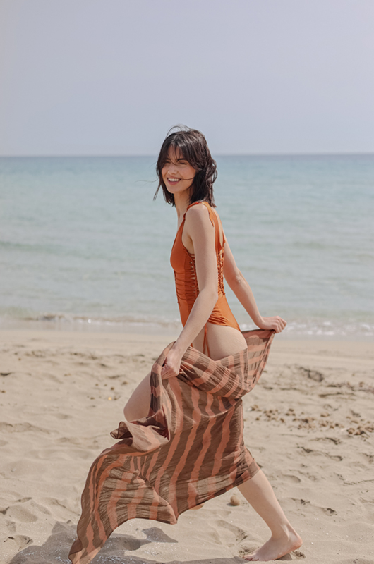 A model walking on the beach wearing an orange one piece swimsuit and a tie dye cotton brown stripes pareo