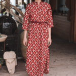 Alt text: Woman standing indoors wearing a long red dress with a geometric pattern, belted at the waist, and teal earrings, posing confidently on a wooden floor.
