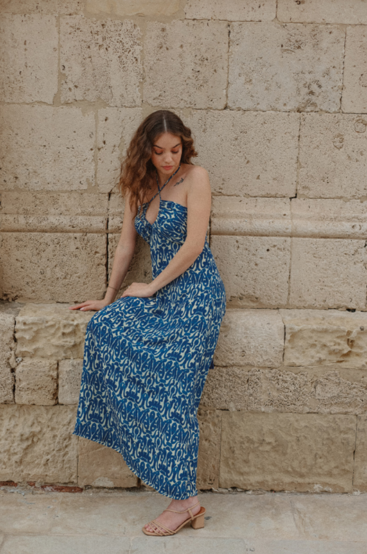 Back view of a woman in a blue and white halterneck maxi dress with an open back and tie detail, highlighting the dress’s rear design and drape.