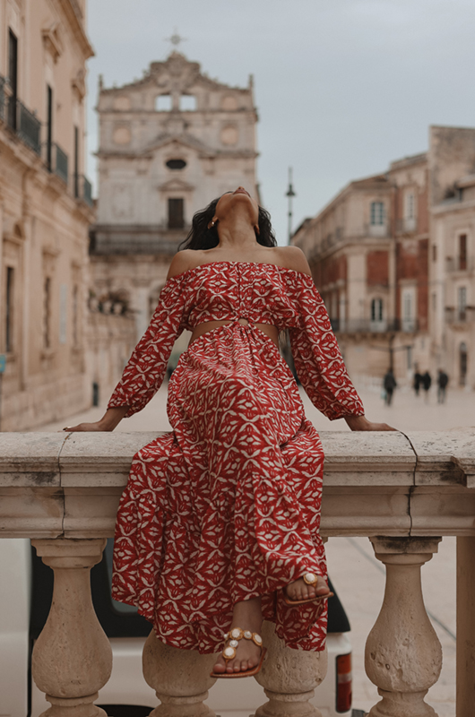 “Woman in red bohemian off-shoulder cutout maxi dress sitting on a stone balcony in historic Italian town”