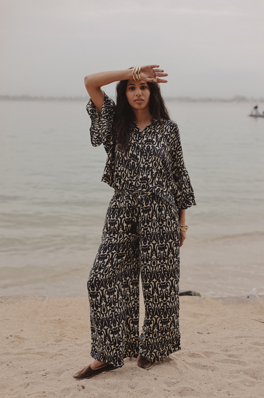 A woman standing on a beach wearing a matching set with a loose patterned shirt and wide-leg pants, holding her arm above her forehead.
