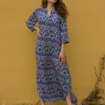 Woman posing outdoors against a mustard yellow wall, wearing a long blue and cream patterned kaftan dress with a front slit and sandals.
