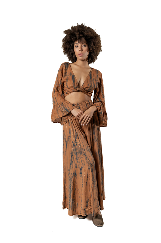 A model poses in a bohemian-style two-piece set with a brown tie-dye pattern. The set includes a cropped top with long, flared sleeves and a twisted knot front, paired with a flowy, high-waisted maxi skirt.