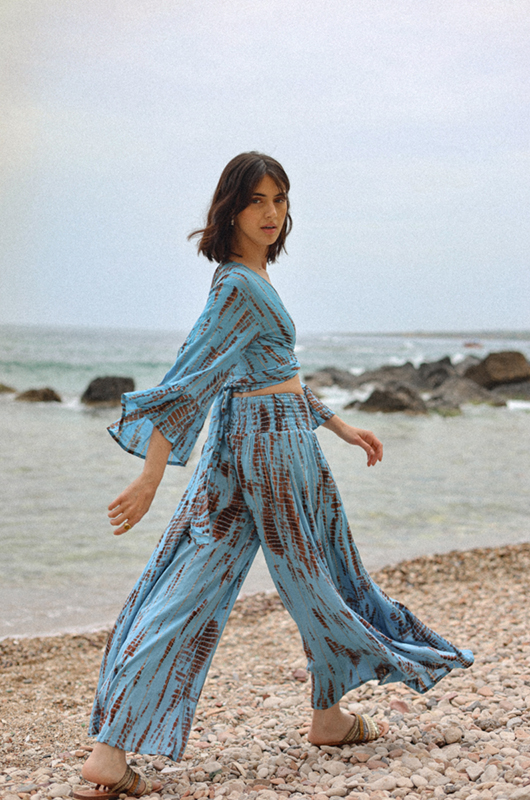 A model walking on a rocky beach in a sky blue and rust tie-dye lurex co-ord set, with wide sleeves and flowy pants.