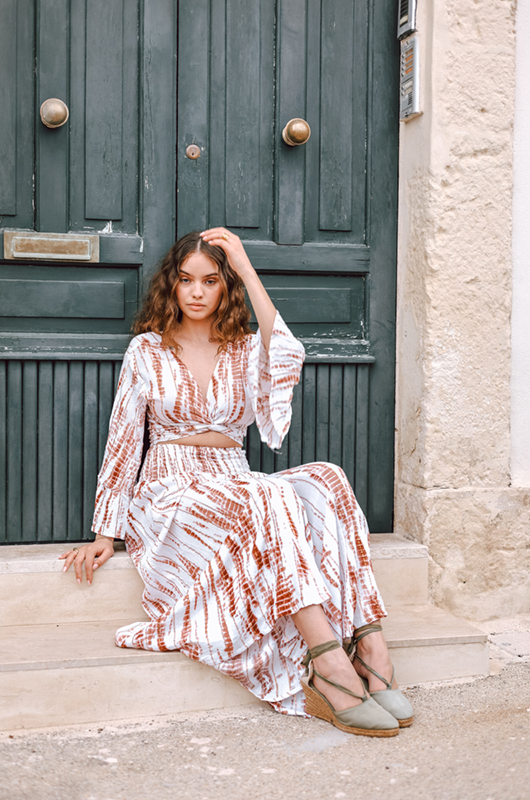 A model sits on stone steps wearing the same white and rust tie-dye lurex set, showing the relaxed bohemian vibe.