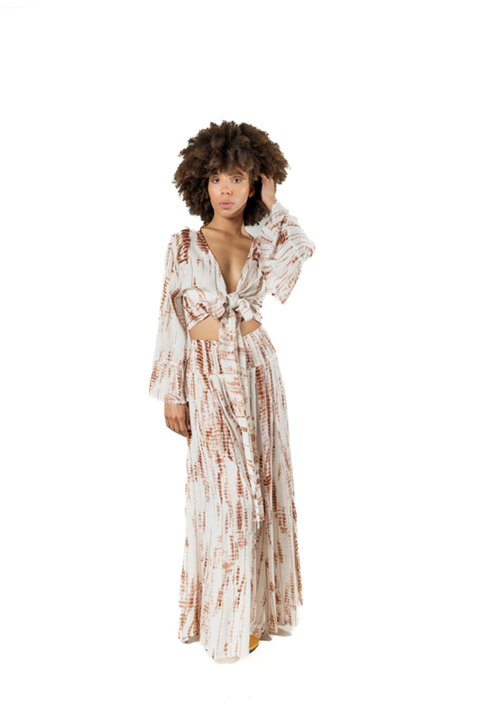 Studio image: A model in a white and rust tie-dye lurex set, standing, with a knotted deep V-neck top and wide-leg trousers.