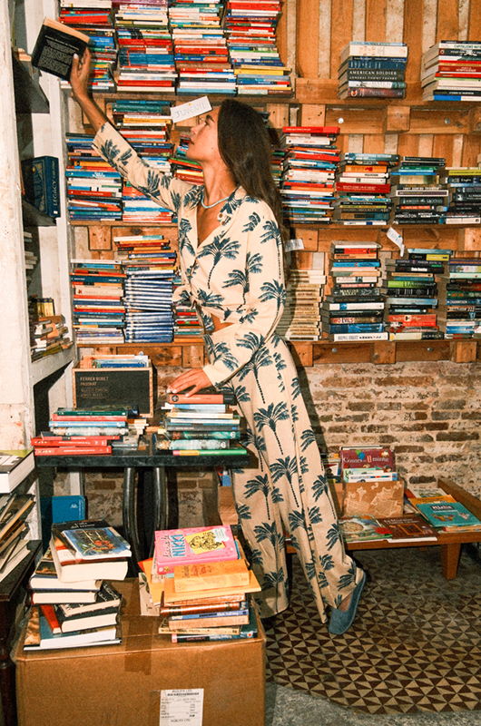 Full-length view of a model reaching for a book in a vintage bookstore, dressed in a matching palm tree print set.