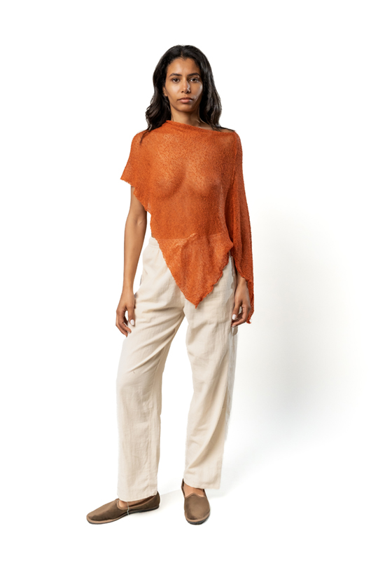 Woman wearing a rust-colored sheer poncho with asymmetrical hem, paired with cream wide-leg pants and brown slip-on shoes, front view.