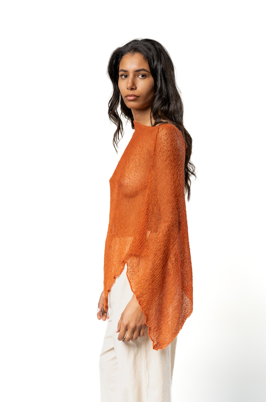 Side view of a woman wearing a rust-colored sheer poncho with an asymmetrical hem, paired with cream wide-leg pants.
