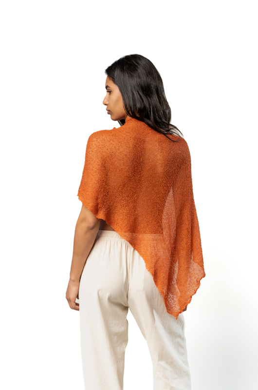 Back view of a rust-colored sheer poncho with an asymmetrical hem worn over cream wide-leg pants.