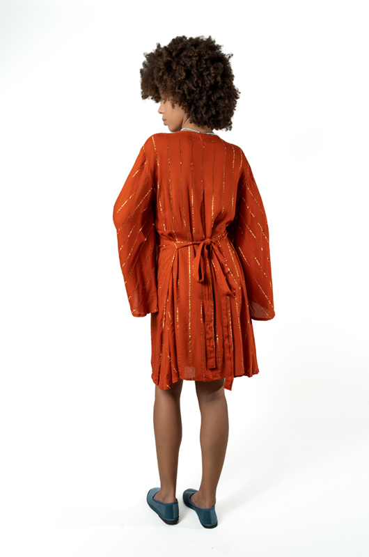 A back view of a woman wearing a rust-orange dress with a tie at the waist and wide bell sleeves, standing with arms relaxed.