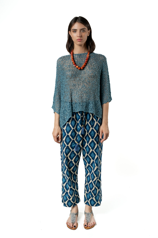 Woman wearing a loose teal open-knit sweater poncho with geometric blue patterned cotton boyfriend trousers and sandals