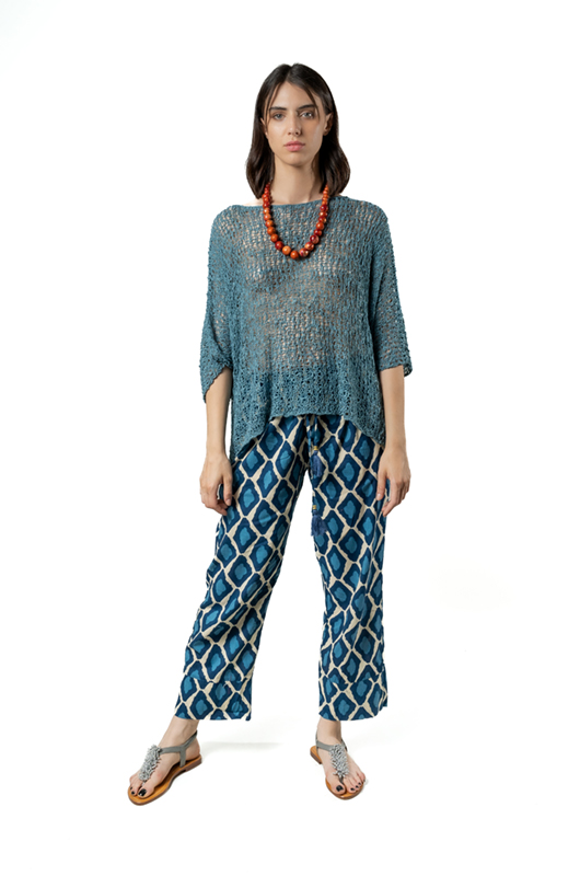 Woman wearing a loose teal open-knit sweater poncho with geometric blue patterned cotton boyfriend trousers and silver sandals, front view