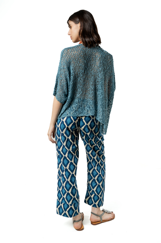 Woman wearing a loose teal open-knit sweater poncho with geometric blue patterned cotton boyfriend trousers and silver sandals, back view