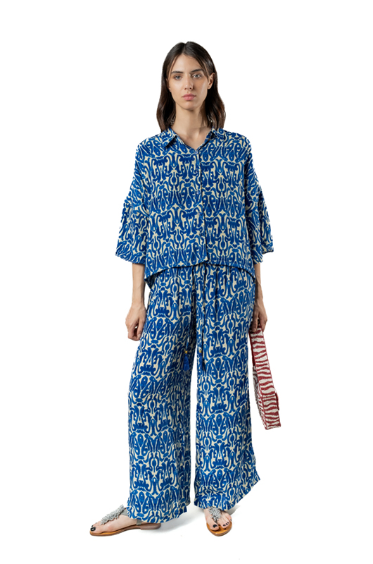 Woman wearing a loose blue and white printed shirt and matching palazzo pants, holding a red patterned bag, studio background.