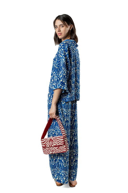 Side view of a woman wearing a blue and white printed co-ord set, holding a red and white patterned handbag, studio background.
