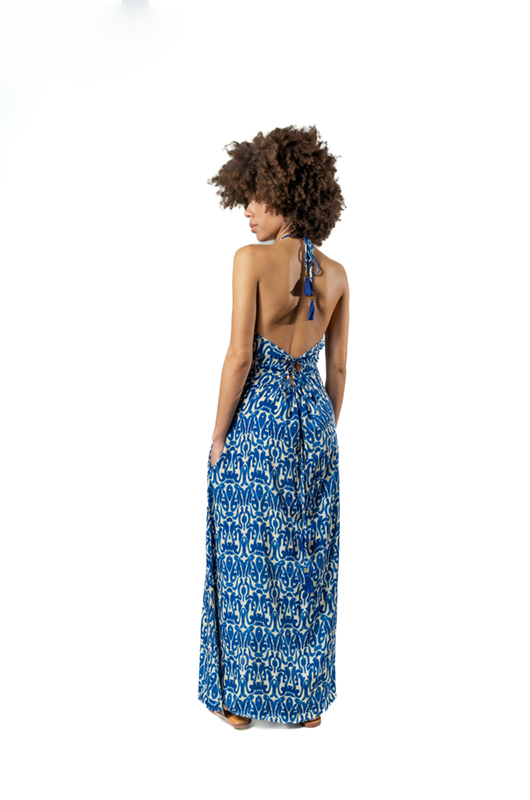 Back view of a woman in a blue and white halterneck maxi dress with an open back and tie detail, highlighting the dress’s rear design and drape.