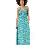 Model facing front in a turquoise halter dress with an abstract white pattern, hands in pockets, on a white studio background.