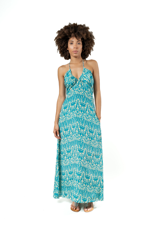 Model facing front in a turquoise halter dress with an abstract white pattern, hands in pockets, on a white studio background.