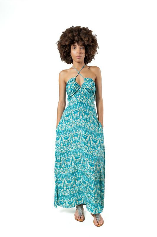 Model facing front in a turquoise halter dress with an abstract white pattern, hands in pockets, on a white studio background.