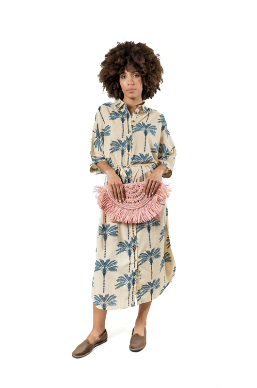Model wearing a beige button-down midi dress with a blue palm tree print, standing next to a vintage blue car, styled with brown flats and a woven clutch.