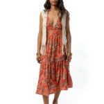 A woman wearing a sleeveless midi dress with a deep V-neckline in an orange tropical floral print, styled with a cream crochet vest and yellow friulana.