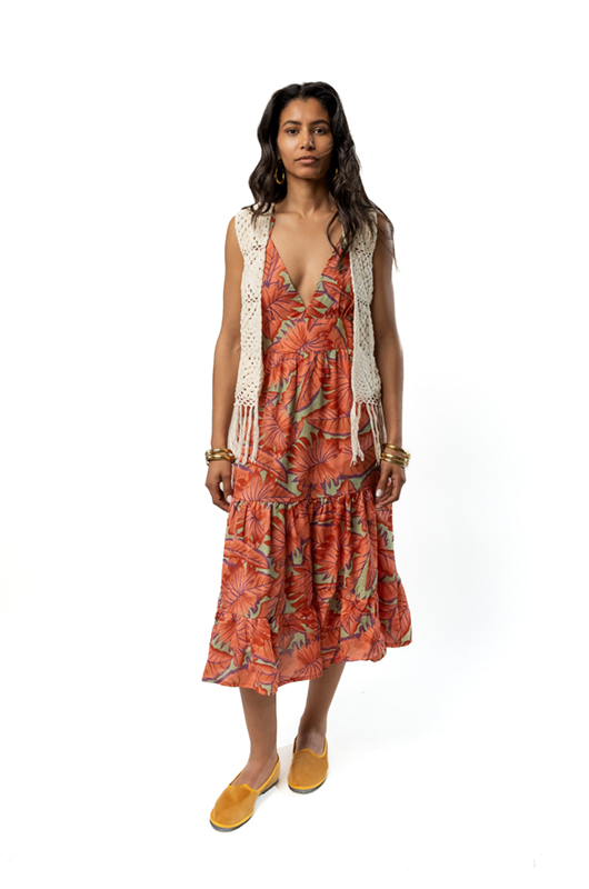 A woman wearing a sleeveless midi dress with a deep V-neckline in an orange tropical floral print, styled with a cream crochet vest and yellow friulana.