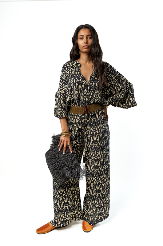 A woman in a patterned outfit, accessorized with a wide belt and holding a fringed bag, posing in a studio.
