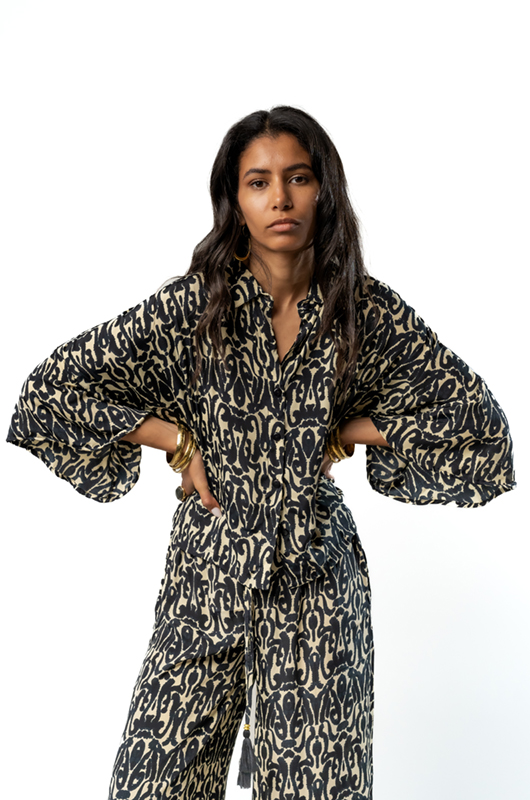 A woman wearing a patterned loose shirt and wide-leg pants posing with hands on her hips.