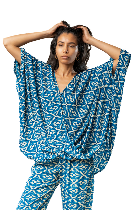 Model wearing a blue patterned oversized draped top with matching pants, posing with arms raised.