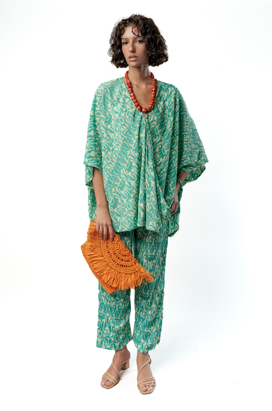 Model wearing a loose green patterned kaftan-style top and pants holding an orange fringed clutch