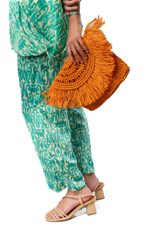Close-up of a model’s hand holding an orange woven clutch, wearing green patterned pants.