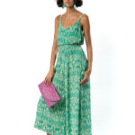 Woman in a green ikat print sleeveless maxi dress with a belted waist, holding a pink clutch and styled with nude sandals.