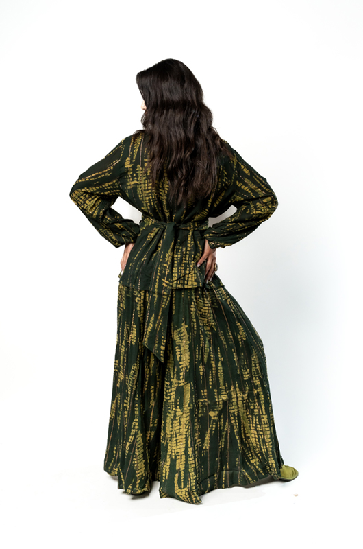 "Back view of a woman wearing a dark green and gold printed kimono-style wrap top with a belt and a matching long skirt."