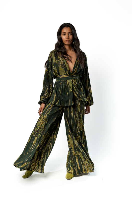 "Action shot of a woman walking, wearing a green and gold printed kimono-style wrap top with a belt and matching wide-leg trousers."