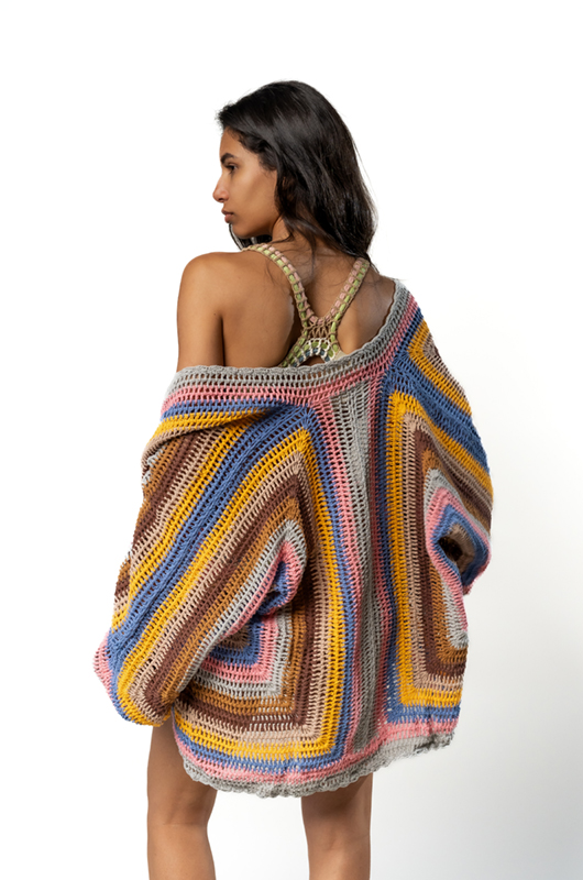 Woman wearing a colorful oversized crochet cardigan, photographed from the back, showing the patchwork pattern of yellow, blue, pink, and brown stripes.