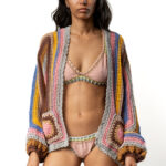Woman in a multicolored crochet cardigan over a light pink crochet-trimmed bikini, standing and looking at the camera.