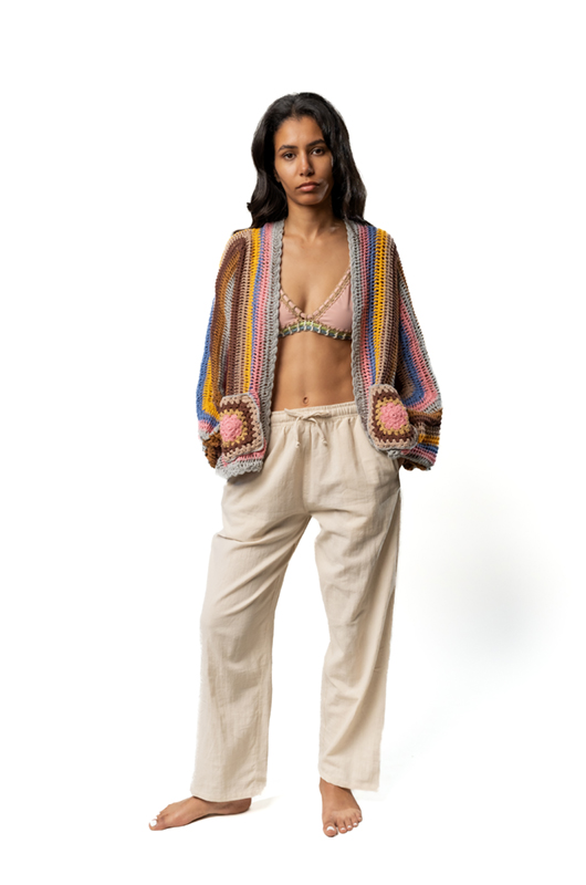Woman wearing a colorful crochet cardigan over a bikini top, paired with loose beige trousers, standing barefoot facing forward.