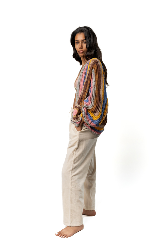 Side view of a woman wearing a crochet cardigan in multicolored stripes with beige trousers, posing barefoot with hands in pockets.