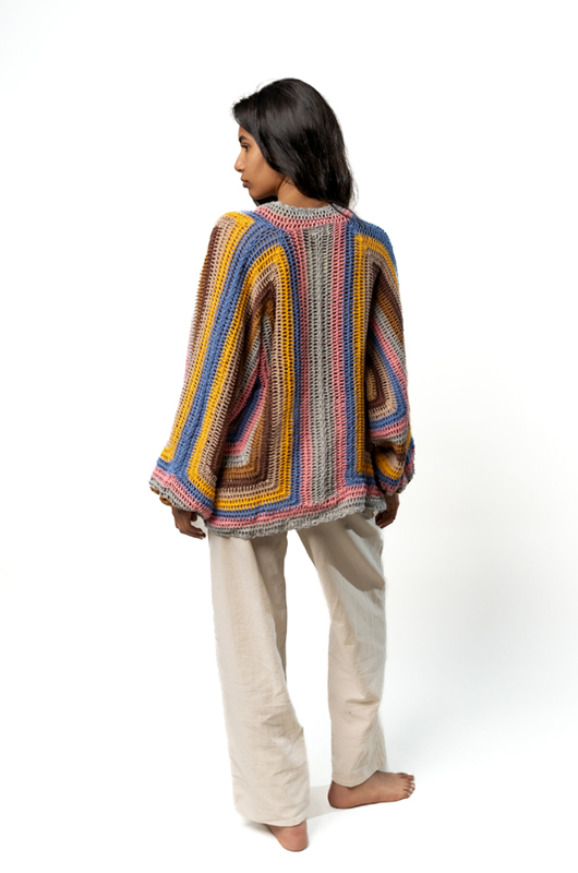 Back view of a woman wearing a vibrant multicolored crochet cardigan with relaxed beige trousers, standing barefoot.