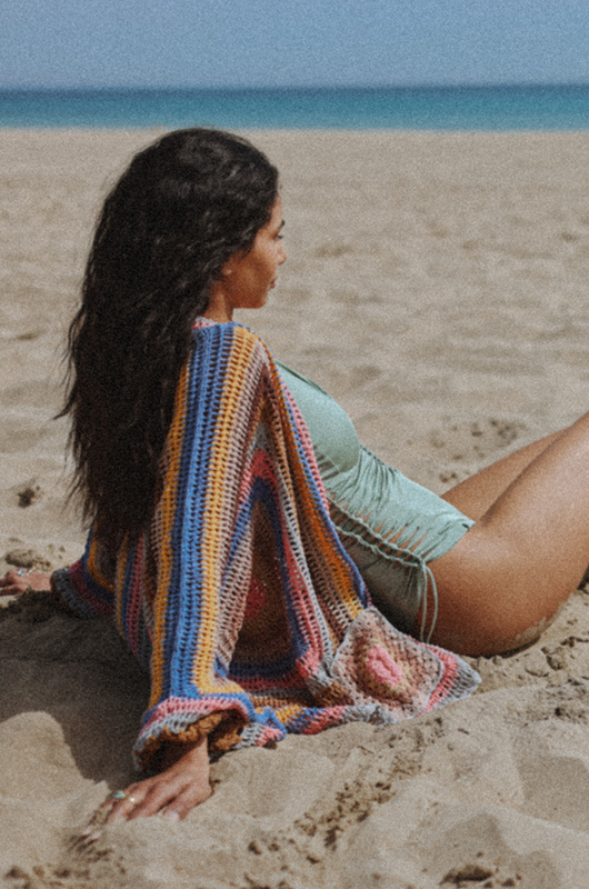 "Smiling woman standing on the beach wearing a sage green swimsuit with a deep neckline and a multicolored crochet cardigan, wind blowing through her hair."