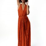Model wearing a floor-length rust-orange halter-neck dress with subtle diagonal gold lurex stripes, tied at the waist, posing with one arm raised.