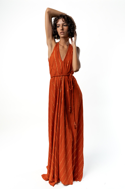 Model wearing a floor-length rust-orange halter-neck dress with subtle diagonal gold lurex stripes, tied at the waist, posing with one arm raised.