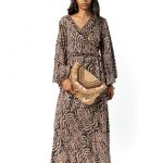 Model wearing a long-sleeved maxi dress with a beige and black abstract leaf pattern, holding a boho-style clutch, front view.