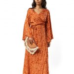 A woman in a long orange patterned wrap dress with flared sleeves, holding a boho-style clutch, posing against a plain background.