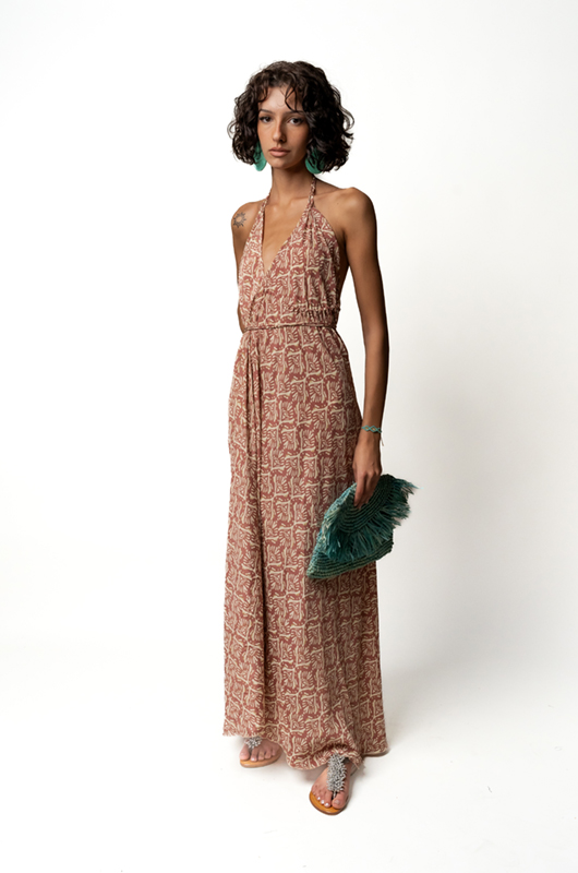 Front view of a woman wearing a halter neck maxi dress with a pink and beige leaf pattern, holding a turquoise fringed clutch."
