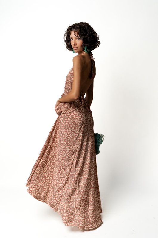 "Three-quarter back view of a woman in a pink and pinkleaf print halter maxi dress, holding a turquoise fringed clutch."