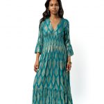 Model wearing a long teal bohemian dress with golden geometric patterns, V-neckline, and flared sleeves, standing facing forward."