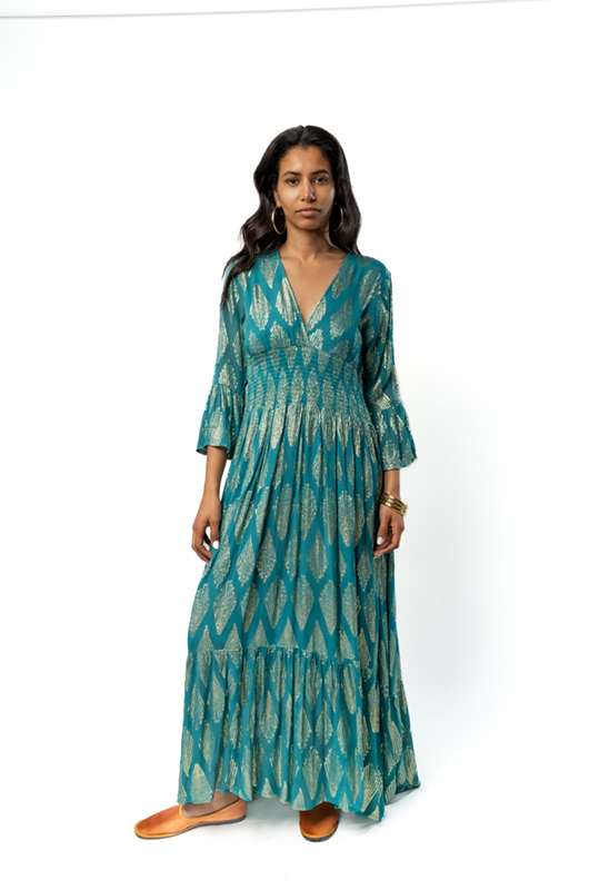 Model wearing a long teal bohemian dress with golden geometric patterns, V-neckline, and flared sleeves, standing facing forward."